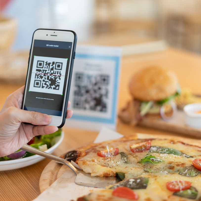 Person scanning a restaurant QR code menu with a smartphone at a table with pizza and a burger.